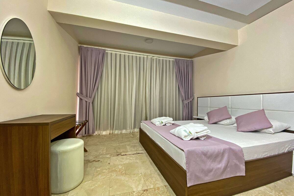 Une chambre d’hôtel avec un lit, une table de chevet en bois, un miroir rond et une salle de bain équipée de toilettes et d’une poubelle.