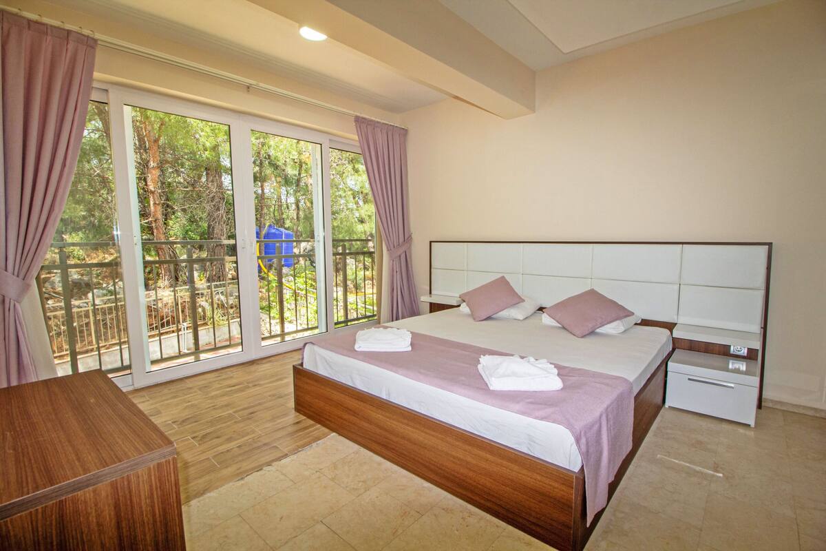 Une chambre avec un grand lit, une table de chevet, une commode en bois et une porte-fenêtre coulissante donnant sur un balcon.