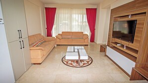 Una sala de estar con un sofá color beige, una mesa de centro, un televisor sobre un mueble de madera y una cortina roja.