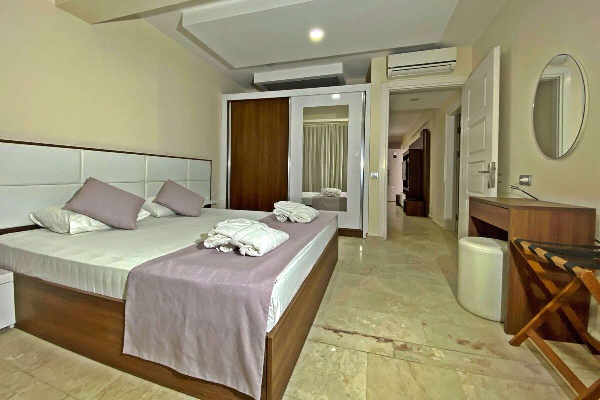 Une chambre d’hôtel avec un grand lit, une tête de lit en bois, une table de chevet, un miroir et une salle de bain visible à travers une porte ouverte.