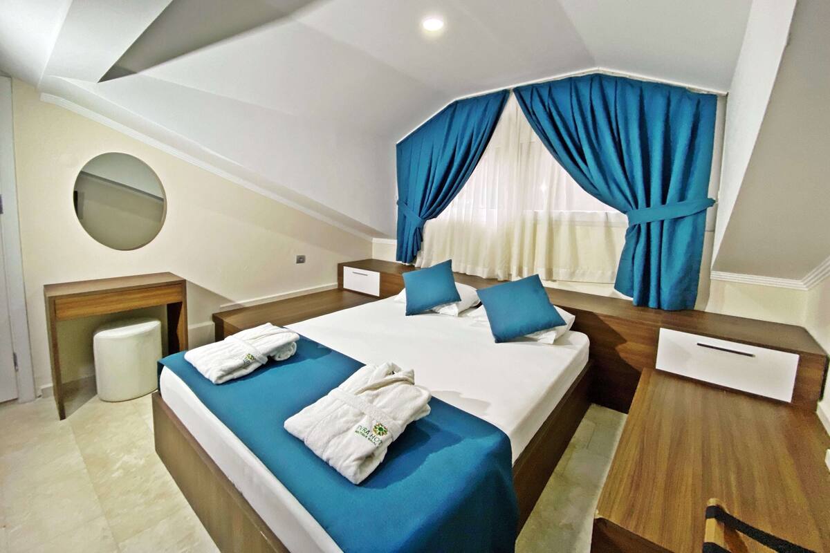 Une chambre d’hôtel moderne avec un grand lit, une literie bleue, un miroir rond, une table de chevet en bois et une vue sur l’extérieur à travers des rideaux transparents.