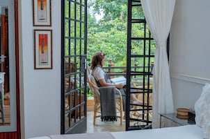 A room with a bed, a chair, a table, and a view of greenery outside.