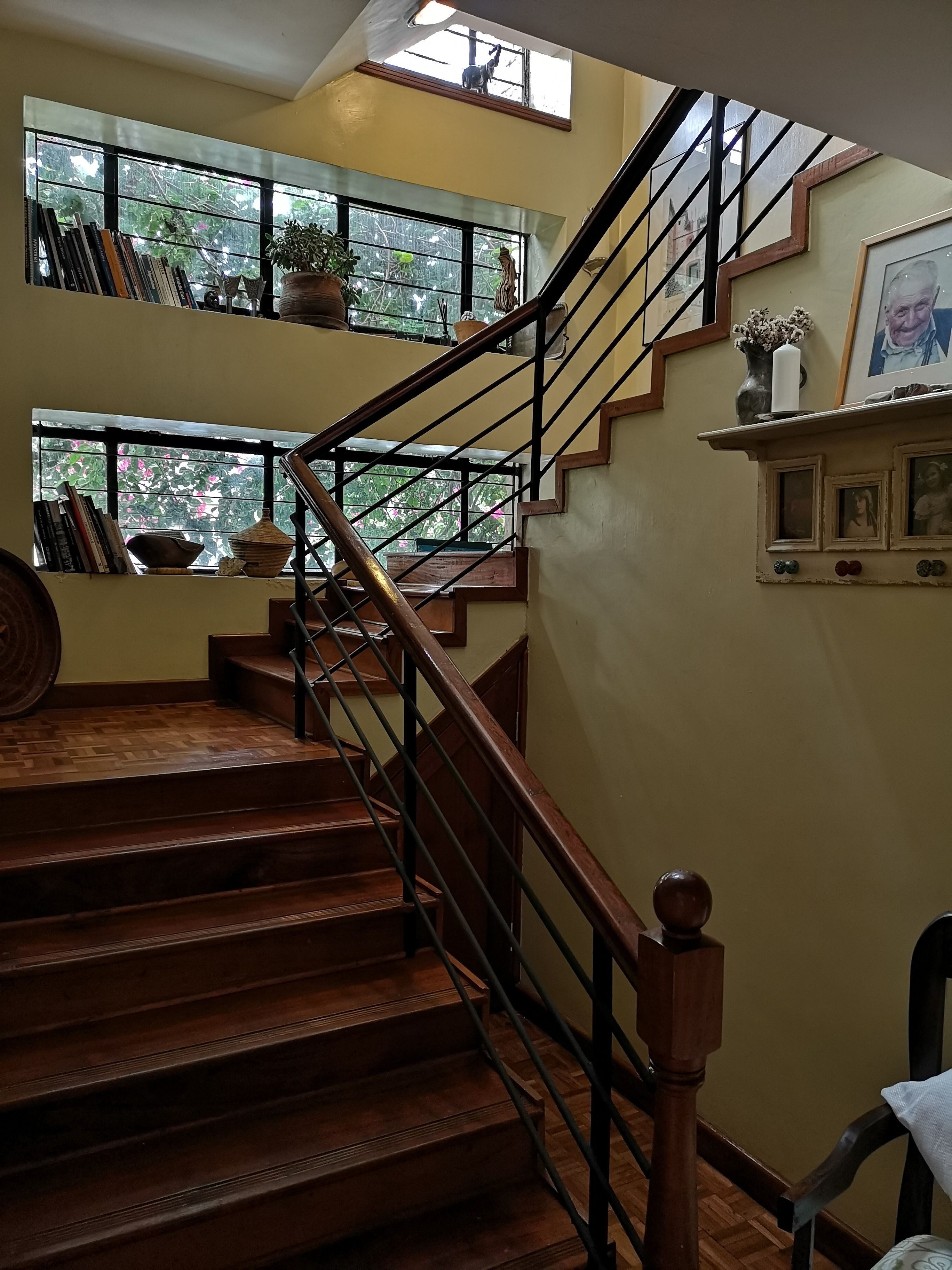 Un escalier en bois, doté d’une rampe en métal, mène à l’étage supérieur. Sur le palier, on trouve des fenêtres, ainsi que des livres et une plante en pot placés sur le rebord de la fenêtre.