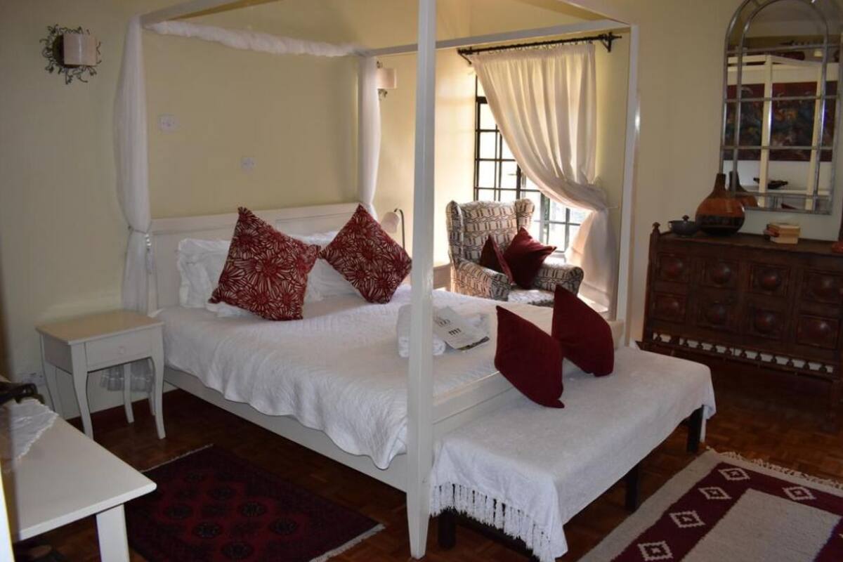 A four-poster canopy bed with white bedding and red pillows, a wooden nightstand, a wicker chair, and a wooden dresser with a mirror.