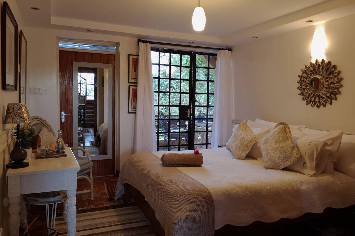 A bedroom with a bed, a desk, a mirror, and a window with a view of greenery.