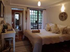 A bedroom with a bed, a desk, a mirror, and a window with a view of greenery.