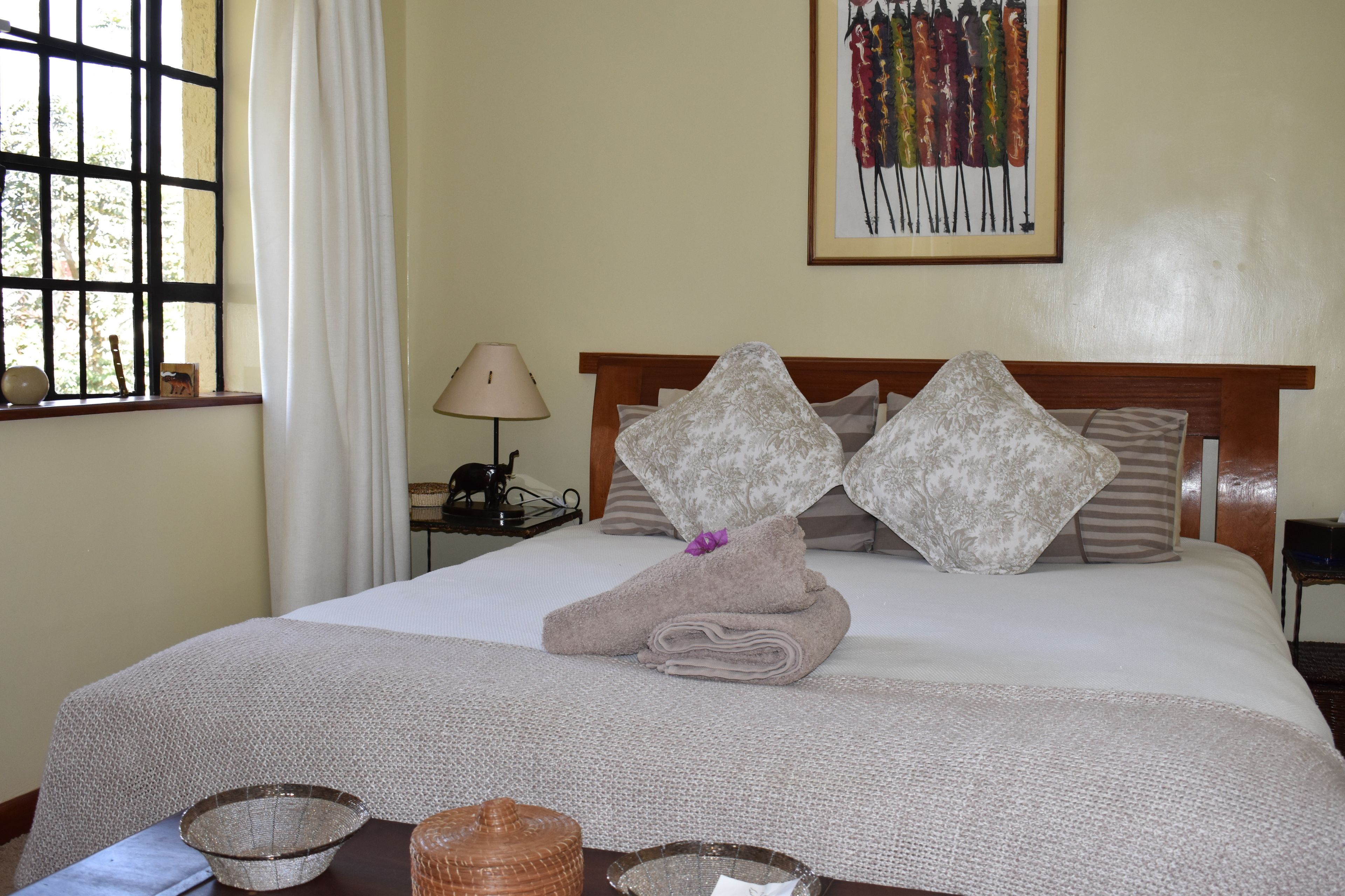 A neatly made bed with a folded towel and two decorative pillows, a lamp, a framed artwork, and a window with a view of trees.