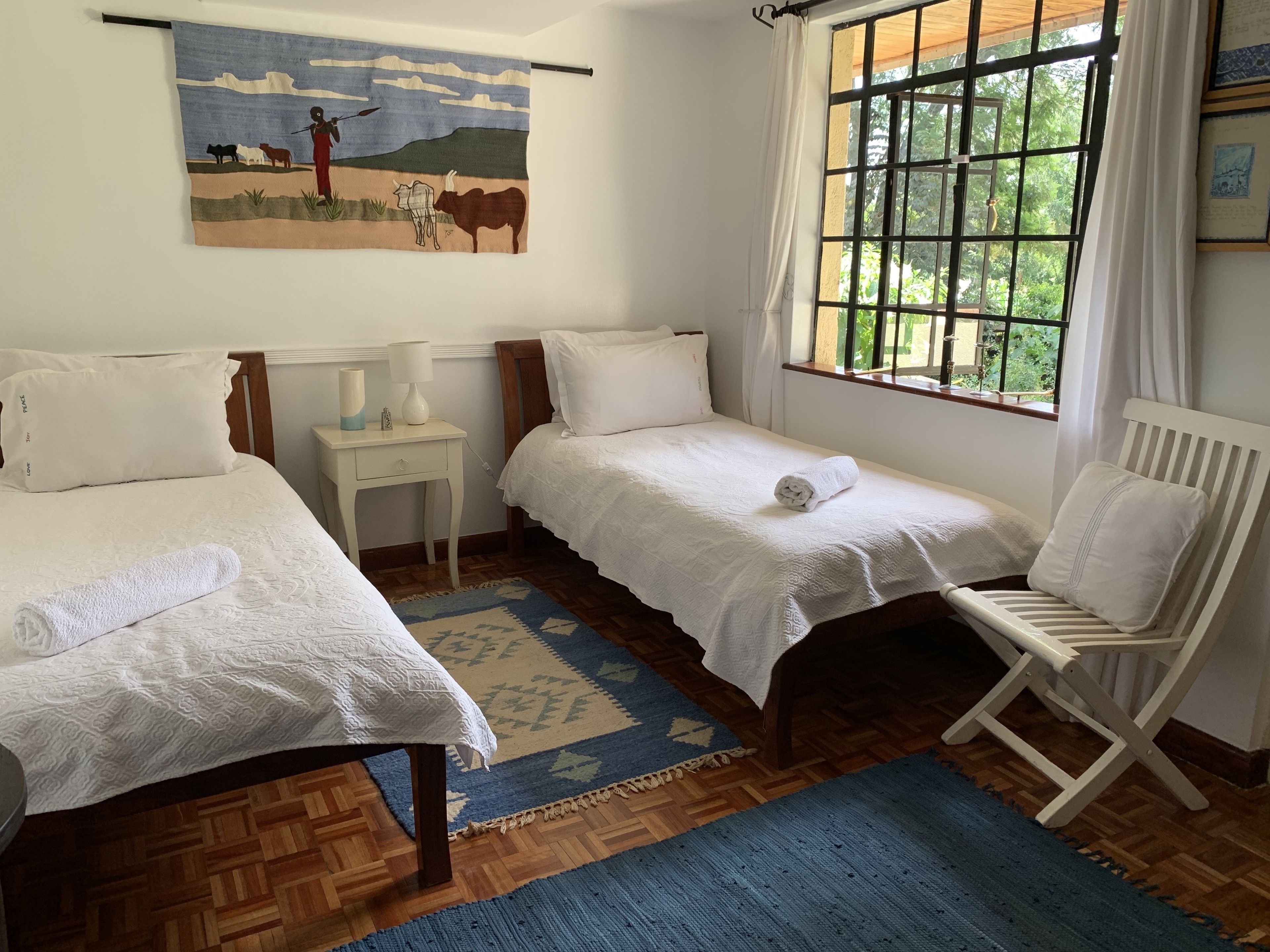 Un dormitorio con dos camas individuales, una mesita de noche, una silla, un ventanal con cortinas y un cuadro en la pared.