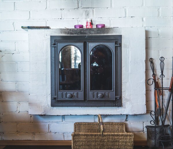 A fireplace with two mirrored doors, a wicker chair in front, and a metal stand with hooks to the right. - Ukonjärven Holiday Village (Inari)