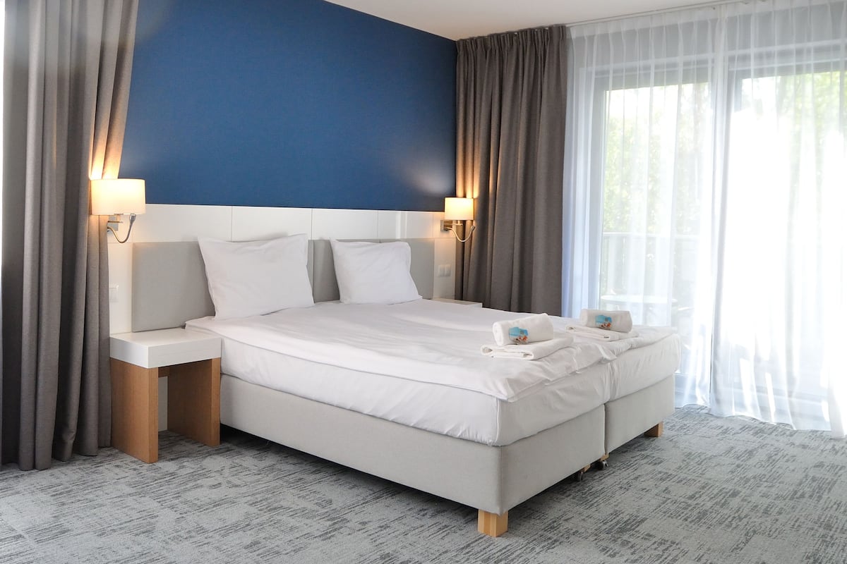 A hotel room with a large bed, two bedside tables, a headboard, and a window with curtains.