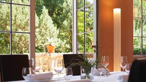 Mesa para dos, con copas de vino y un jarrón de flores, ventanas amplias con vistas a la vegetación y una cálida fuente de luz en la pared.