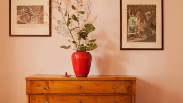 Un aparador de madera con tres cajones y un jarrón rojo con plantas encima. Dos cuadros están colgados en la pared sobre el aparador.