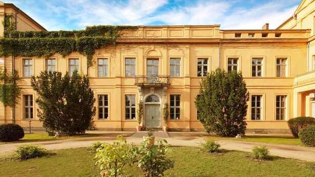 Un edificio amplio y amarillo con una fachada simétrica, una entrada central con un arco decorativo y un jardín bien cuidado al frente.