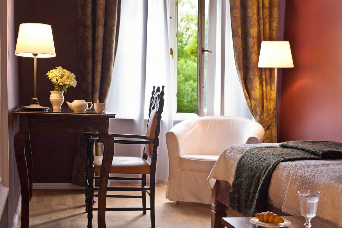 A hotel room with a bed, a chair, a desk with a lamp, a vase with flowers, and a view of greenery outside.