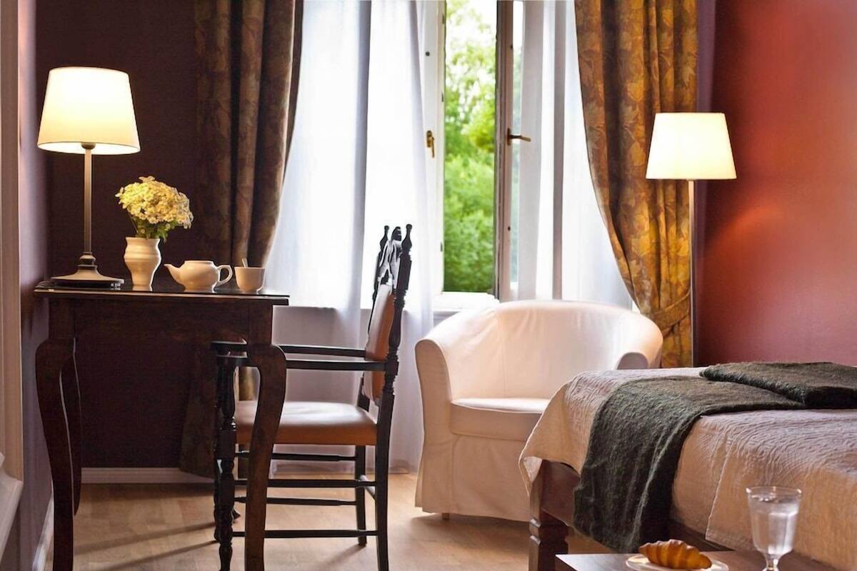 A hotel room with a bed, a chair, a desk with a lamp, a vase with flowers, and a window with curtains.