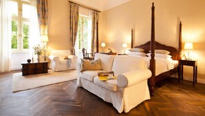 Un dormitorio espacioso con una cama balinesa grande, una zona de estar con sofá y sillones, y un piso de madera con diseño de espiga.
