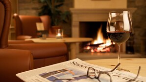 A glass of red wine on a table with a newspaper and glasses, in a warmly lit room with leather seating and a fireplace. - Allgäuer Kräuteralm (Oberstaufen)