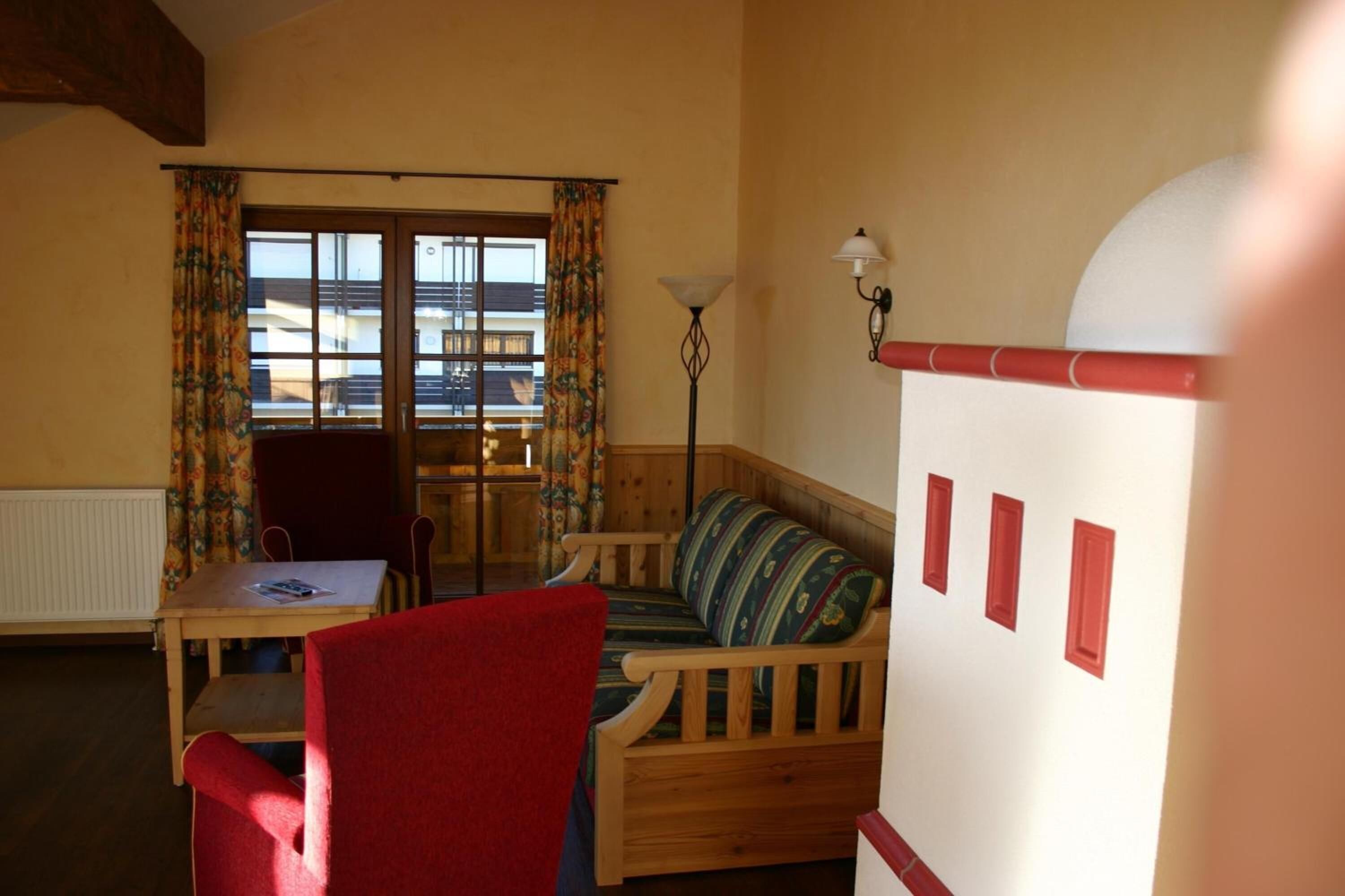 Une pièce chaleureuse avec un canapé en bois, un fauteuil rouge, une table avec une lampe et une fenêtre avec des rideaux.