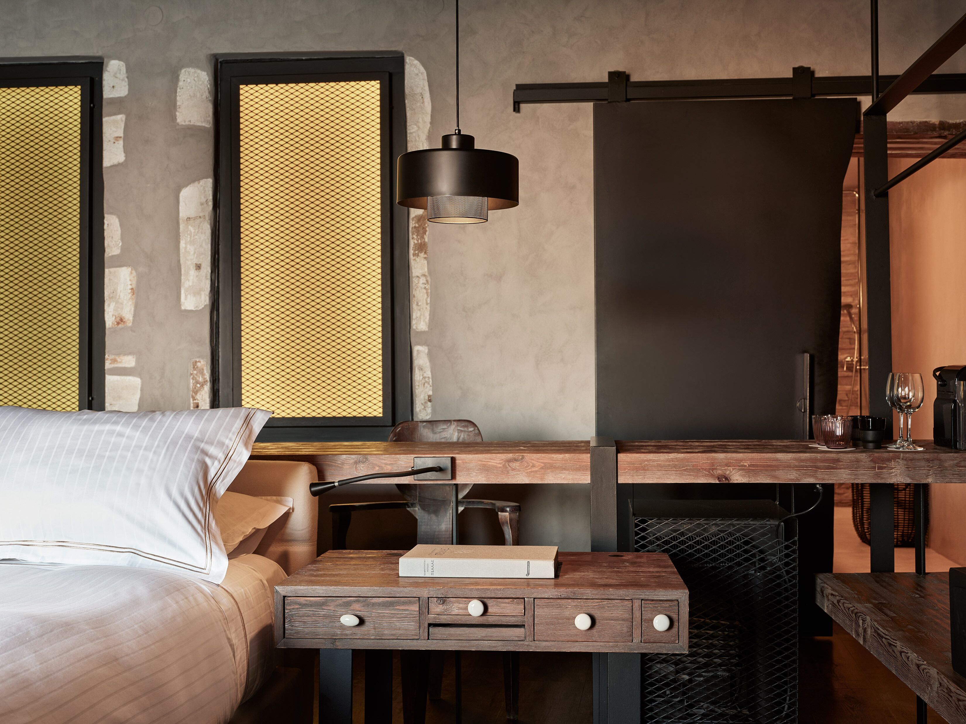 A modern hotel room with a bed, a wooden desk, a hanging light, and a large window with a textured view.