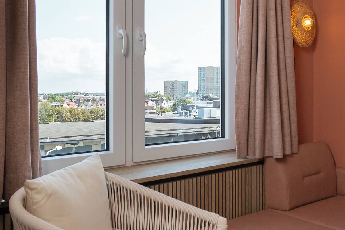 A room with a view of a cityscape through two windows, a wicker chair, a cushioned bench, and a wall-mounted light fixture.