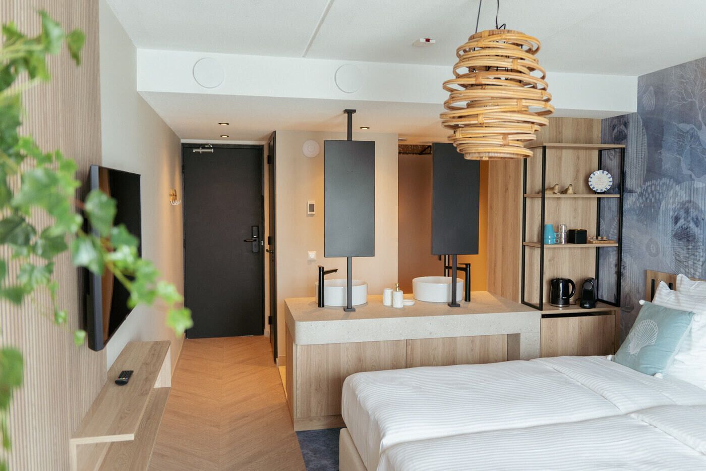 Ein modernes Hotelzimmer mit Holzboden, einem Bett mit weißer Bettwäsche, einem Holztisch mit Spiegel, einem an der Wand montierten Fernseher und einer Korbschlafzimmerlampe.