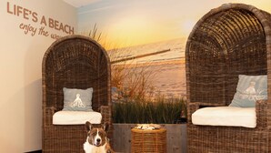 Una habitación con dos sillones de mimbre con cojines, un mural en la pared con una playa, y un perro sentado adelante.