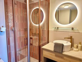 Un baño moderno con ducha envuelta en vidrio, un espejo redondo y un mostrador color beige con lavamanos y artículos de aseo.