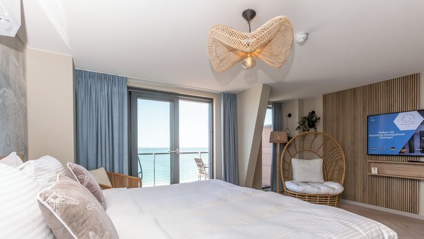Una habitación de hotel con una cama grande, televisión, una silla de mimbre y vista al mar.