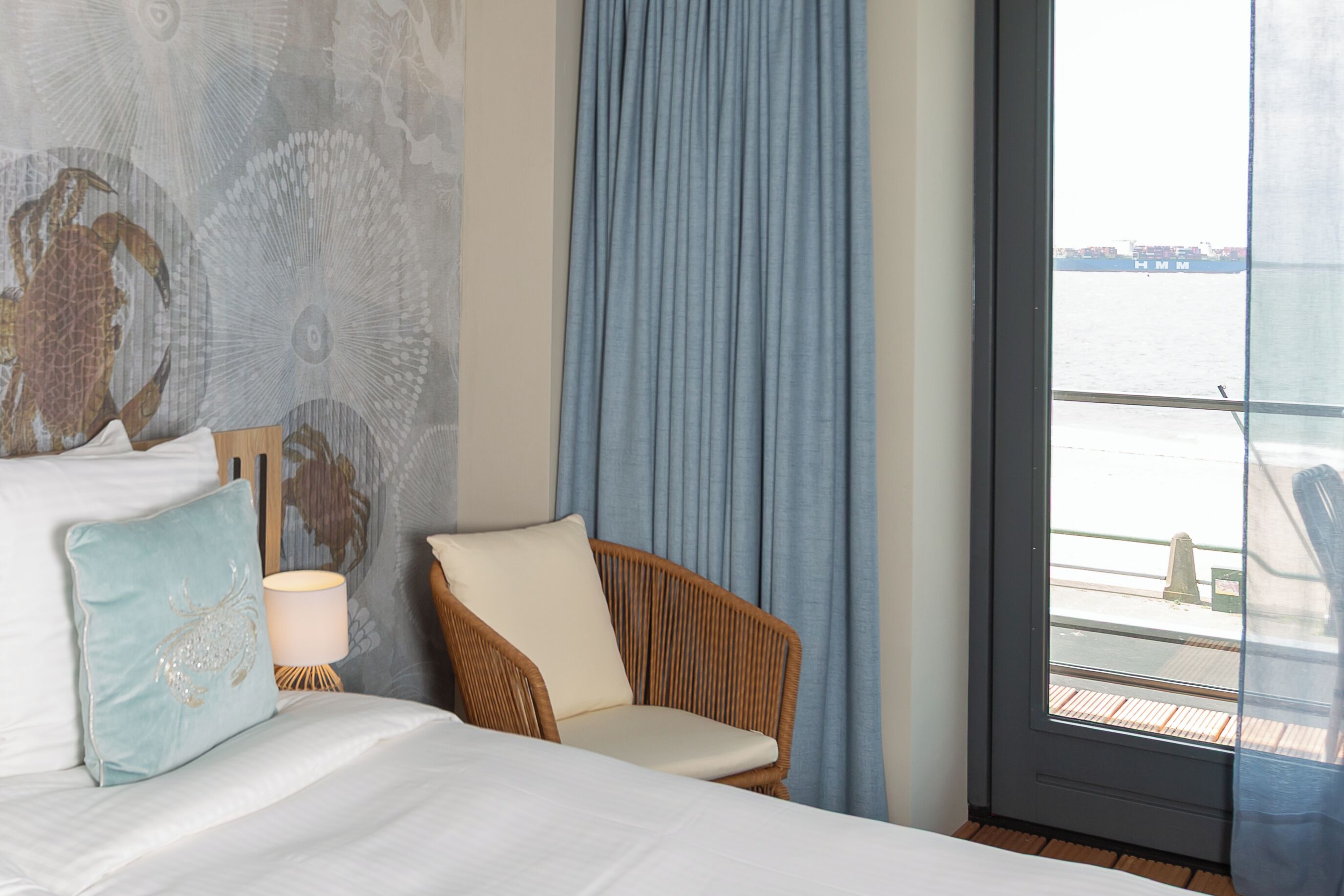 A hotel room with a bed, a wicker chair, a nightstand with a lamp, and a view of the water through a window.