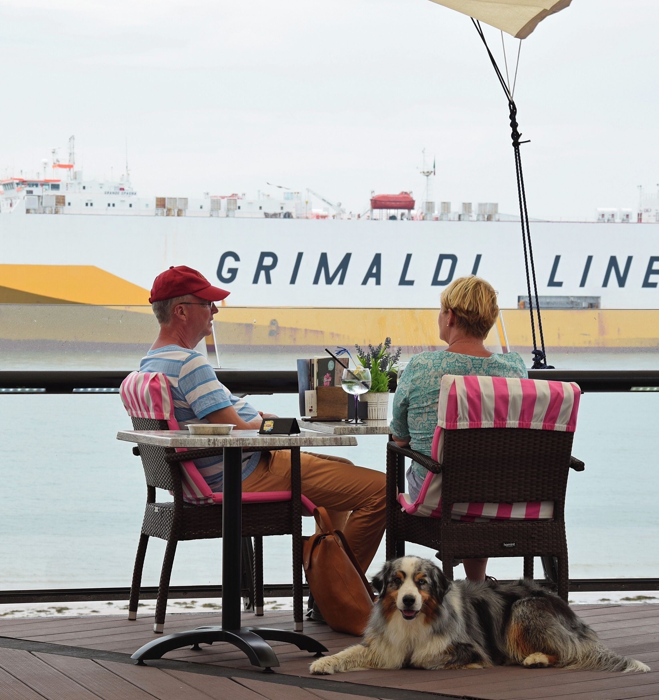 Two people seated at a table with a view of a large ship named 'GRIMALDI LINE' in the background.