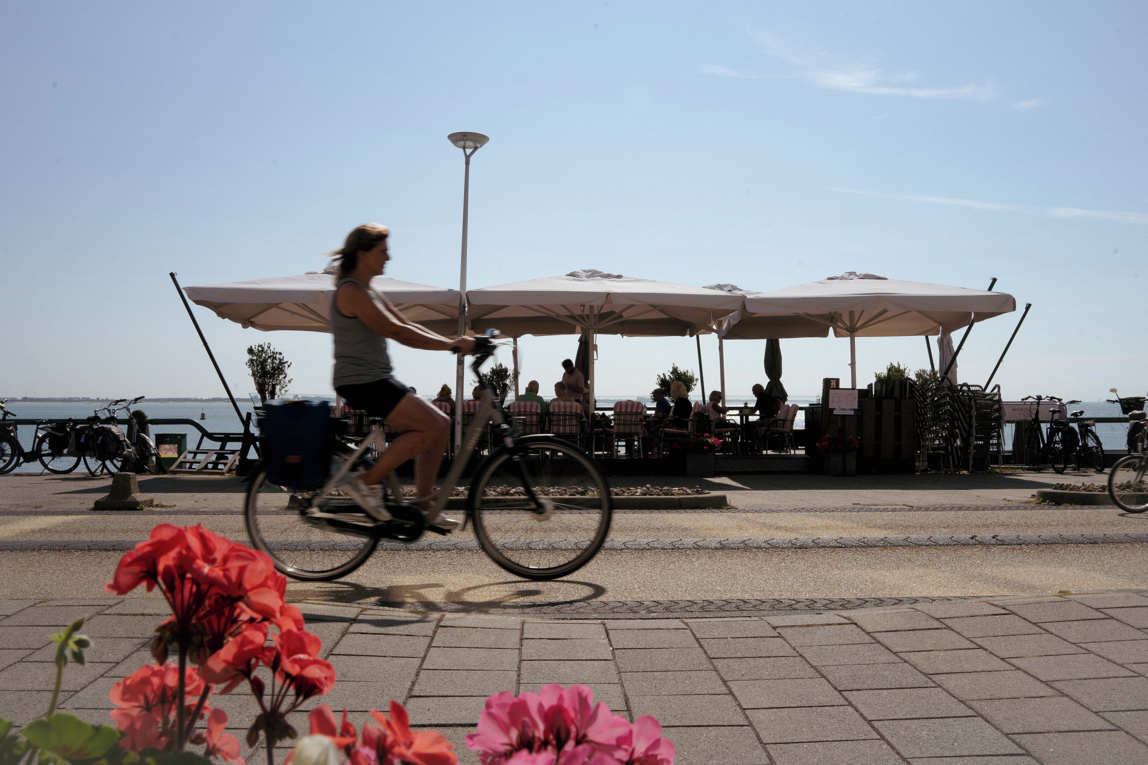 Une personne à vélo circule sur une zone pavée, à proximité d’un espace extérieur aménagé avec des sièges et des parasols.