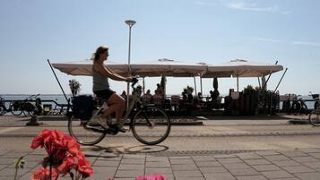 Une personne à vélo circule sur une zone pavée, à proximité d’un espace extérieur aménagé avec des sièges et des parasols.