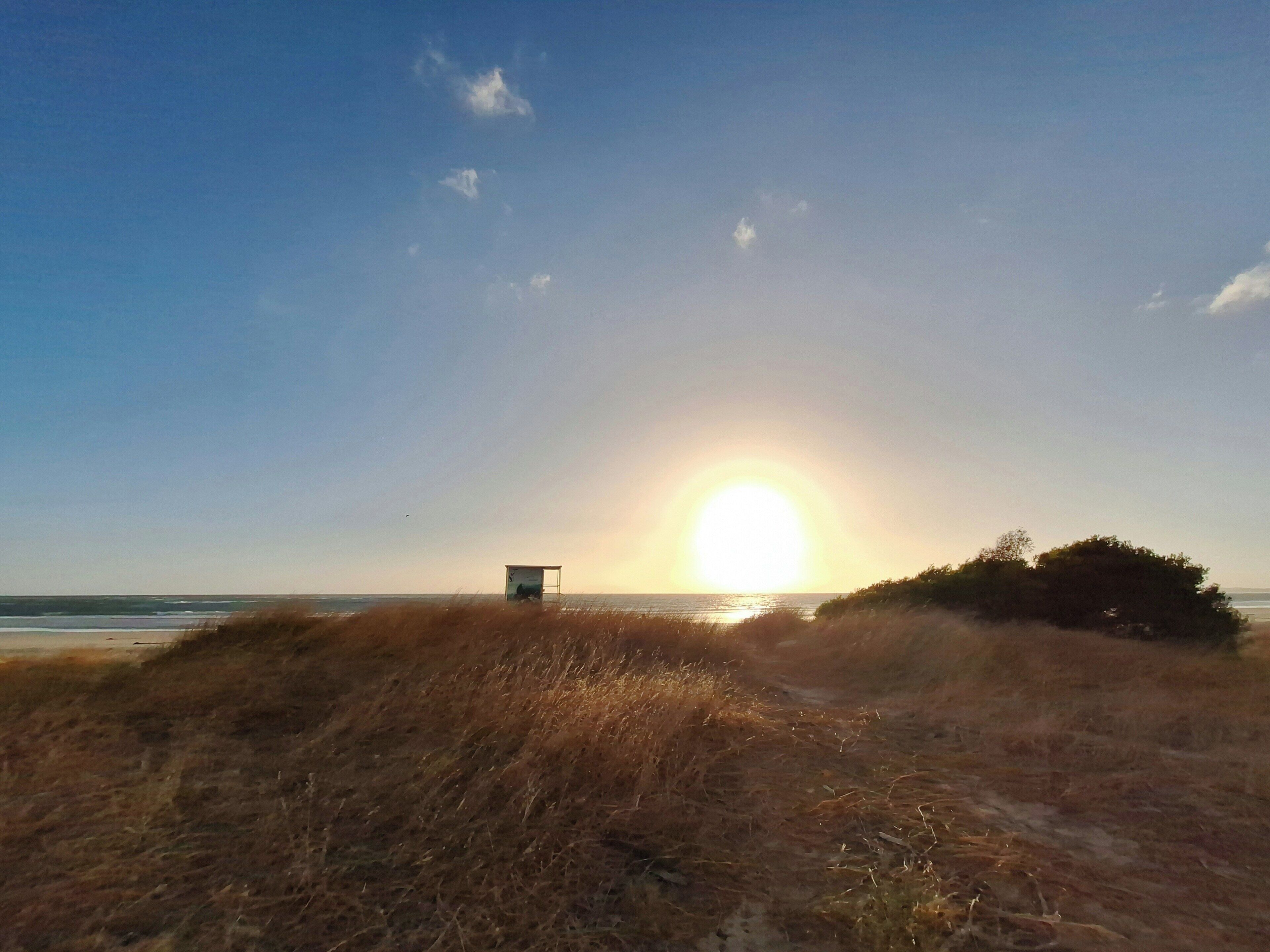 Una spiaggia con una duna, una piccola costruzione e il sole che tramonta sull'oceano.