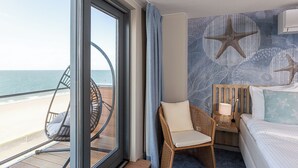 Habitación de hotel con un ventanal grande con vistas a la playa, una silla de mimbre, una cama con un cojín estampado con estrellas de mar y un mural de estrellas de mar.