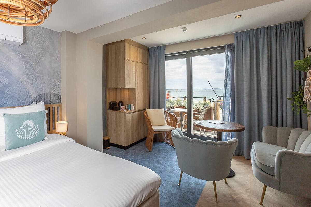 A hotel room with a bed, a seating area with a table and chairs, a kitchenette, and a balcony with a view of the sea.