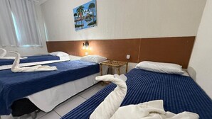 A hotel room with two beds, each with a blue bedspread and white pillows. There is a painting on the wall above the beds, a small wooden table, and a swan-shaped towel on the floor. - pousada sal e mar (ipojuca)