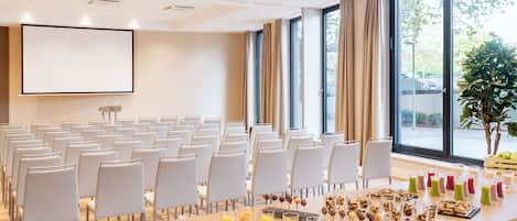 A conference room with rows of white chairs, a large table with various food items and drinks, and a projector screen on the wall.