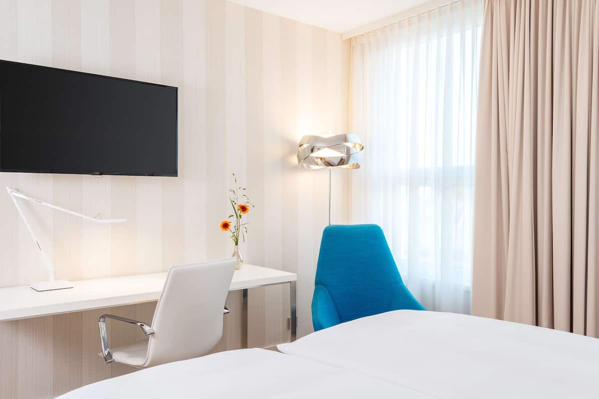 A hotel room with a bed, a desk with a chair, a flat-screen TV, a desk lamp, a vase with flowers, and a window with curtains.