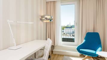 Habitación de hotel moderna con un escritorio blanco, un sillón azul, una lámpara de pie y un ventanal con cortinas.