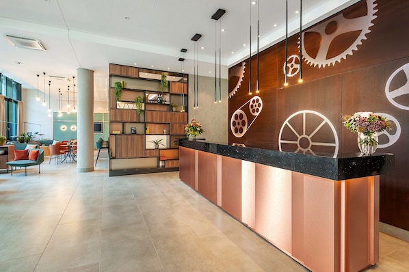Un espace d’accueil moderne avec un comptoir en bois, des étagères ornées de plantes et un grand mur décoré de motifs représentant des engrenages.