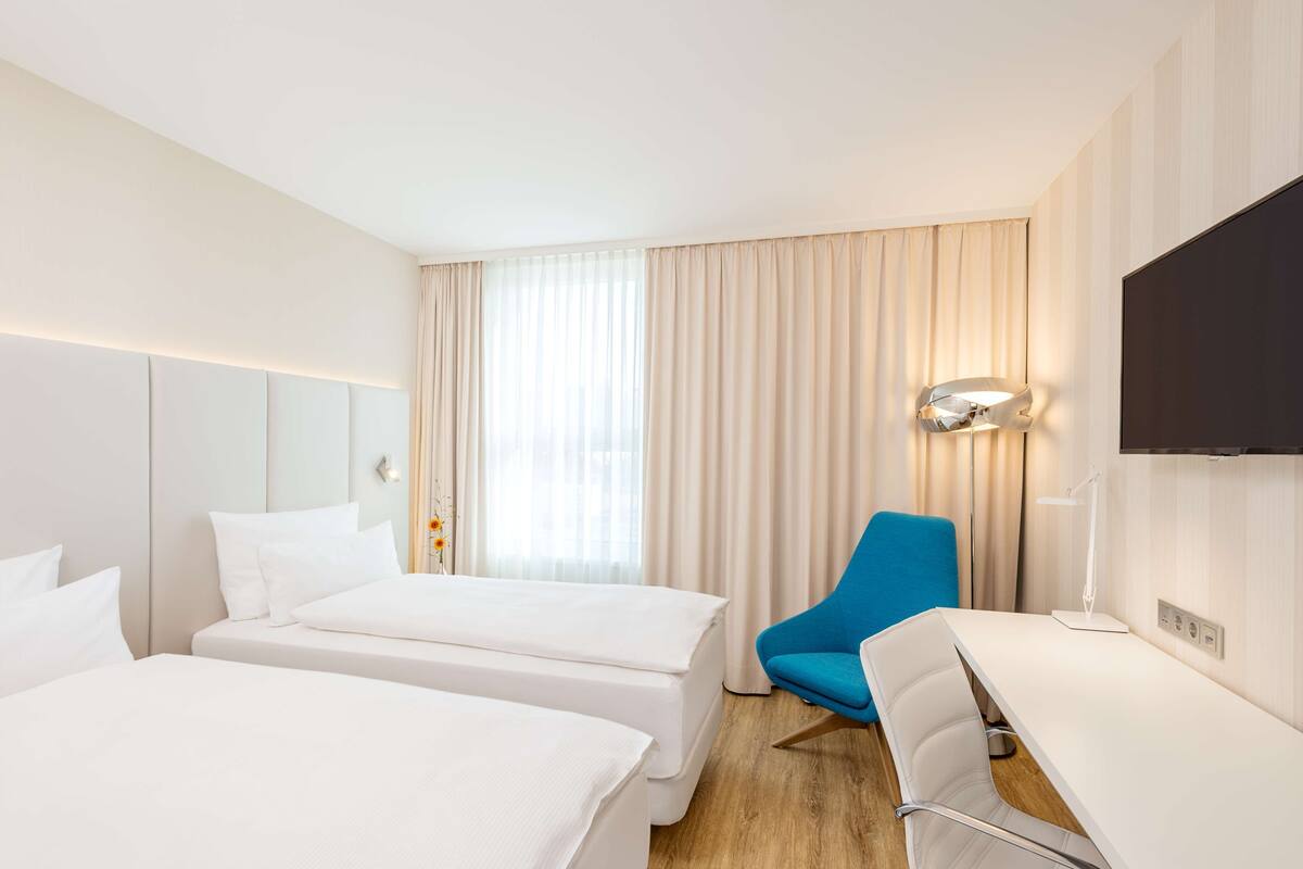 A hotel room with two beds, a desk, a chair, a TV, and a window with curtains.