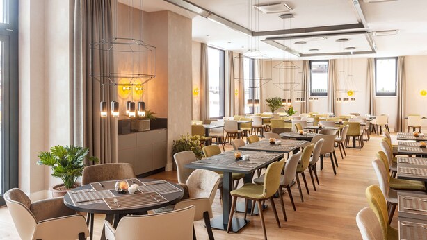 Un vaste espace de restauration d’hôtel, doté de nombreuses tables et chaises, de grandes fenêtres et d’un éclairage moderne.