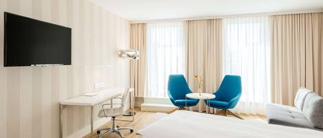 Habitación de hotel con una cama grande, un escritorio con televisor de pantalla plana, dos sillas azules alrededor de una mesita y un sofá.