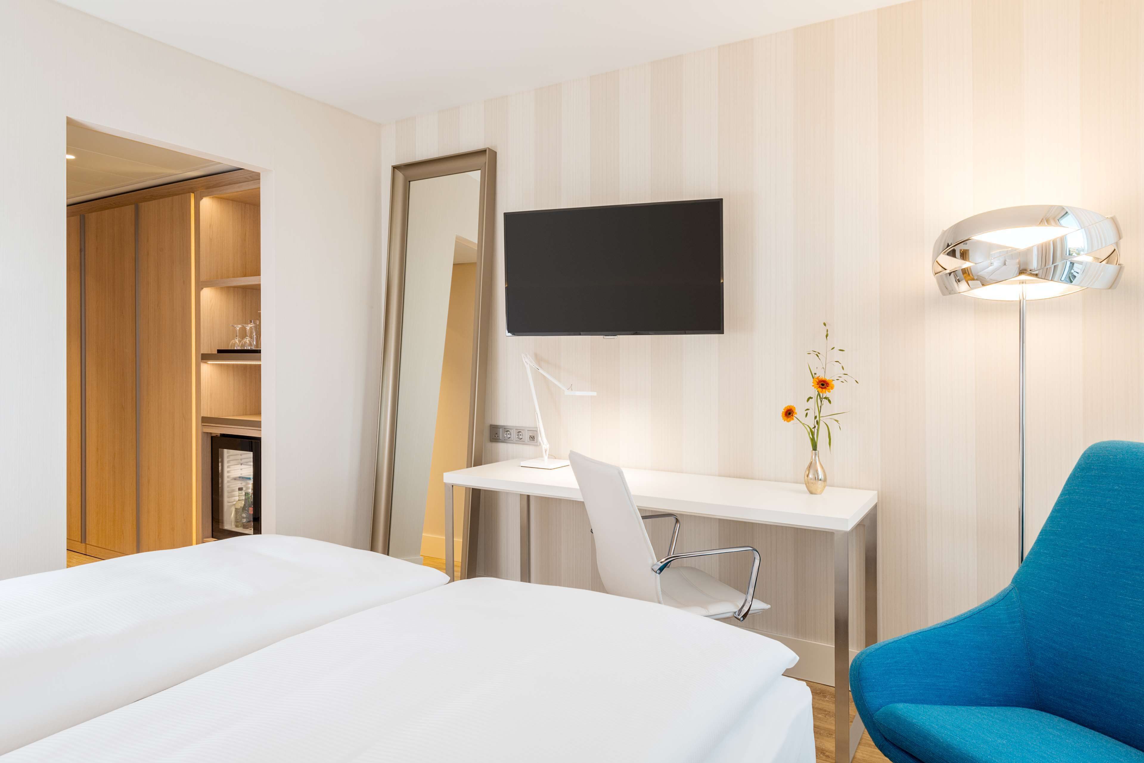 Habitación de hotel con una cama, un escritorio con silla, un televisor, una lámpara y un florero con flores.
