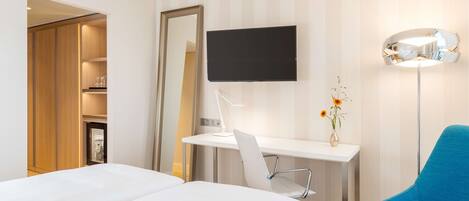 Habitación de hotel con una cama, un escritorio con silla, un televisor, una lámpara y un florero con flores.