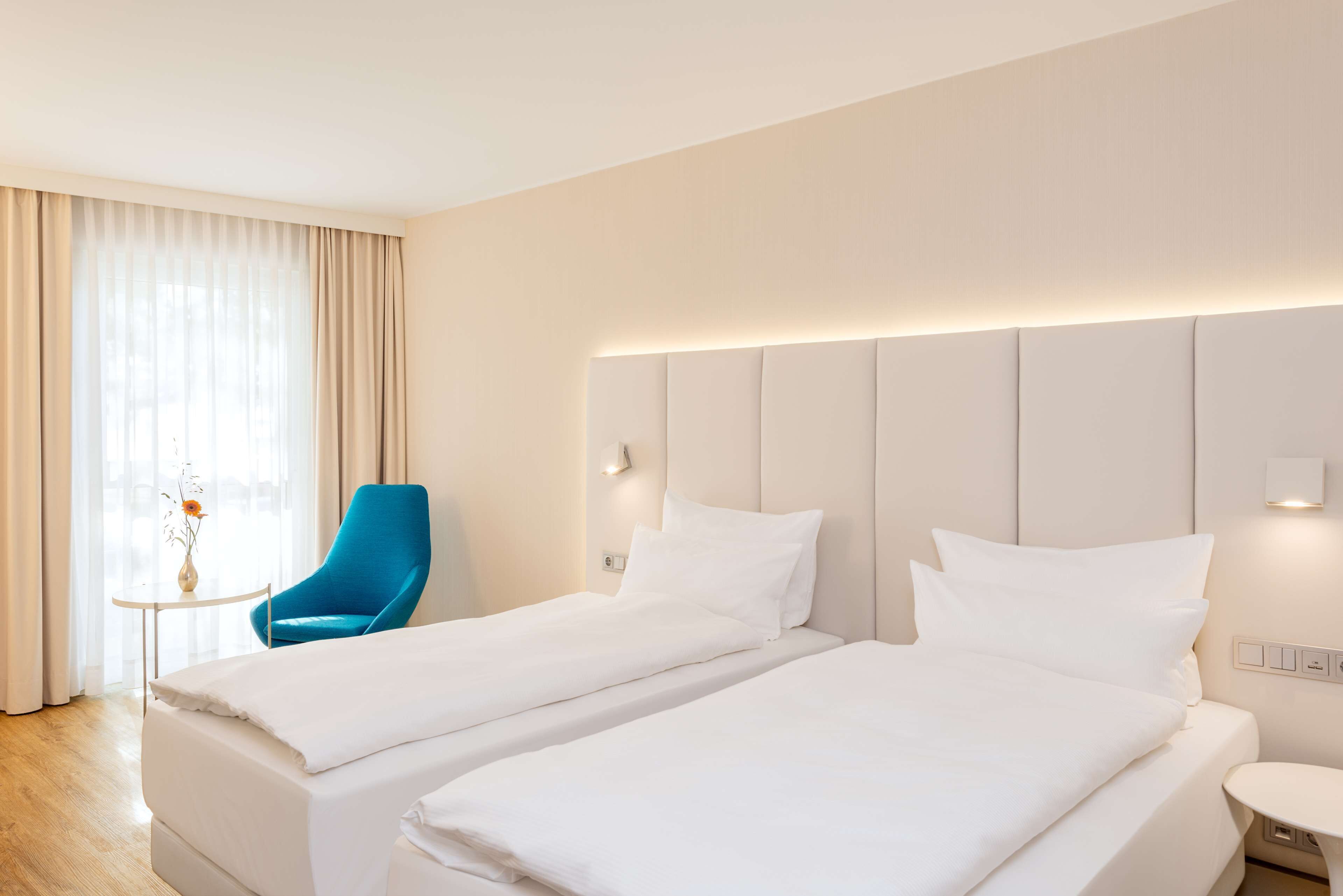 Ein Hotelzimmer mit zwei Einzelbetten, einem blauen Sessel, einem kleinen Tisch und einem großen Fenster mit Vorhängen.
