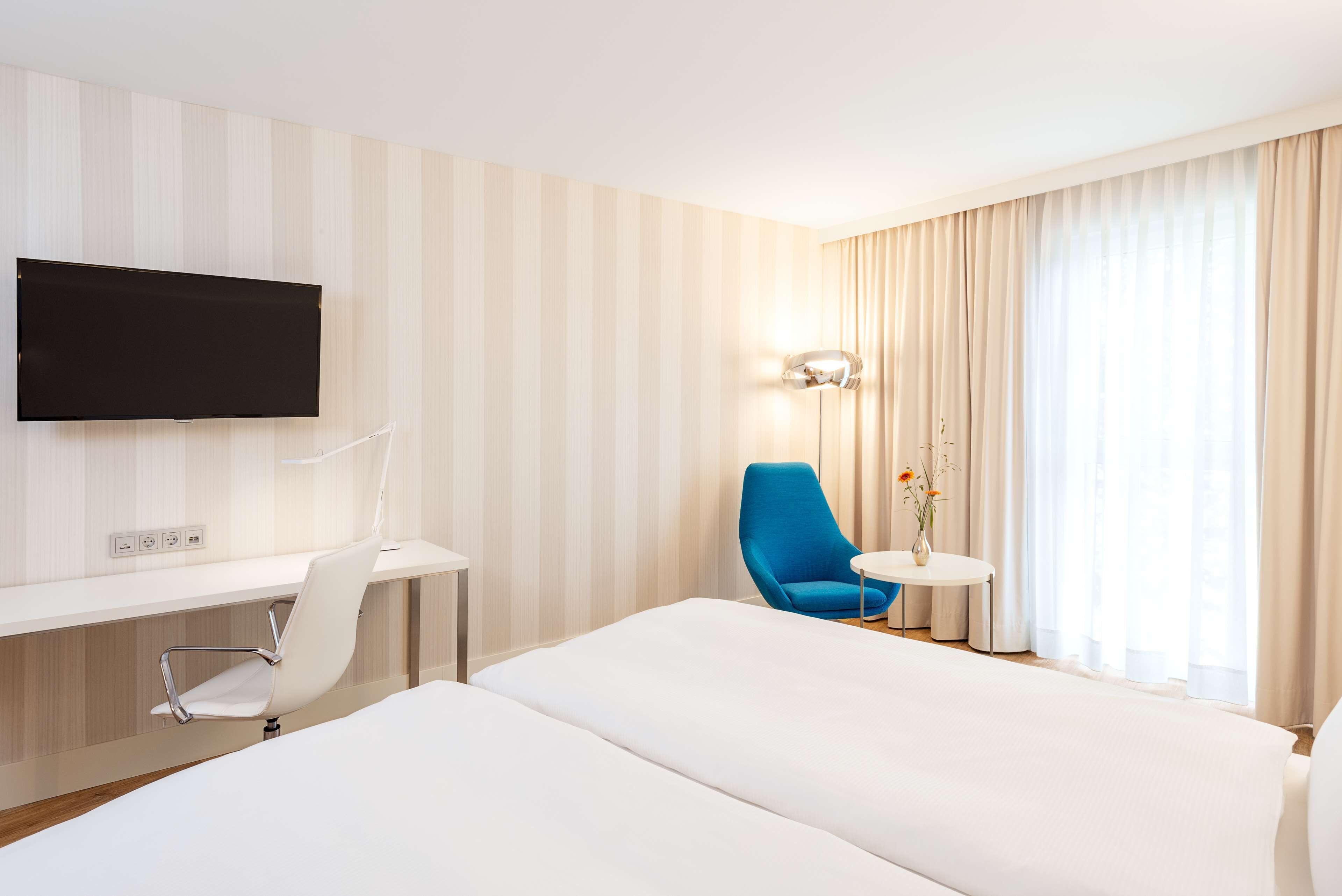 A hotel room with a bed, a desk, a chair, a TV, a lamp, and a window with curtains.