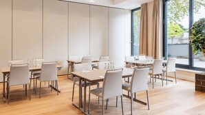 Une salle de conférence moderne, dotée de plusieurs tables et chaises, de grandes fenêtres avec rideaux et offrant une vue sur l’extérieur.