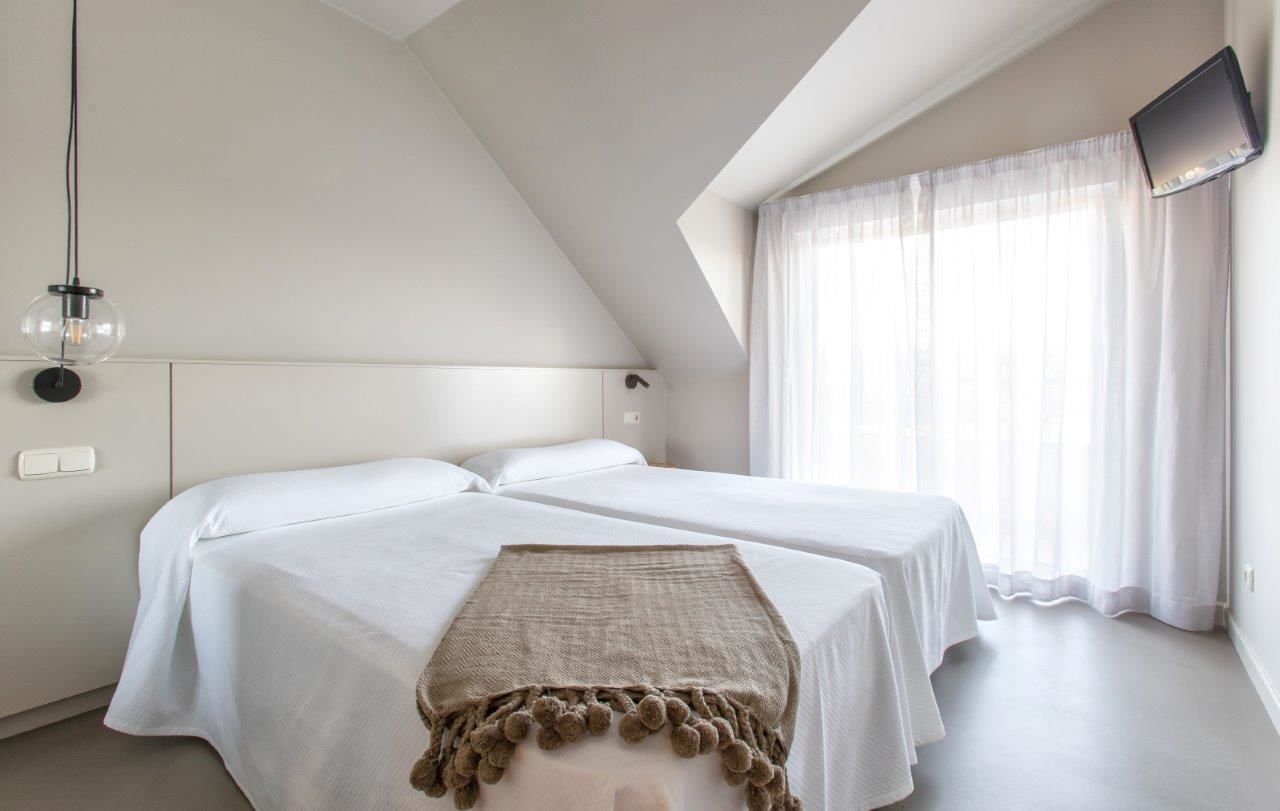 A bedroom with two beds, a TV mounted on the wall, and a large window with sheer curtains.