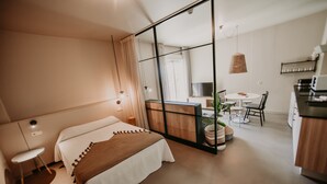 A modern hotel room with a bed, a small desk, a chair, a dining table, and a kitchen area. - Apartamentos Dena (Meano)